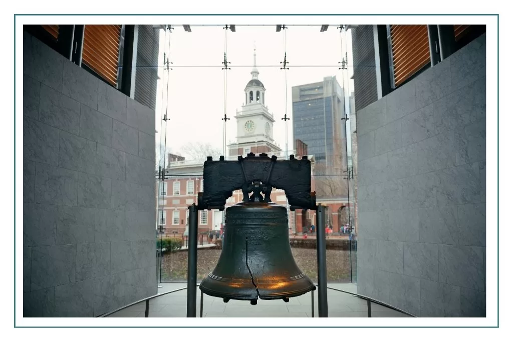 A view of the Liberty Bell on display in Philadelphia with Independence Hall through the window behind it.