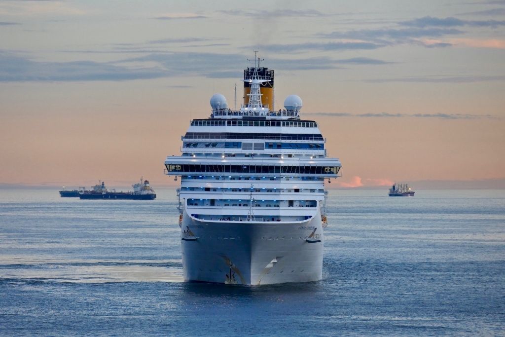 A cruise ship on the ocean sailing towards the camera with other ships and an orange and blue sunset in the background