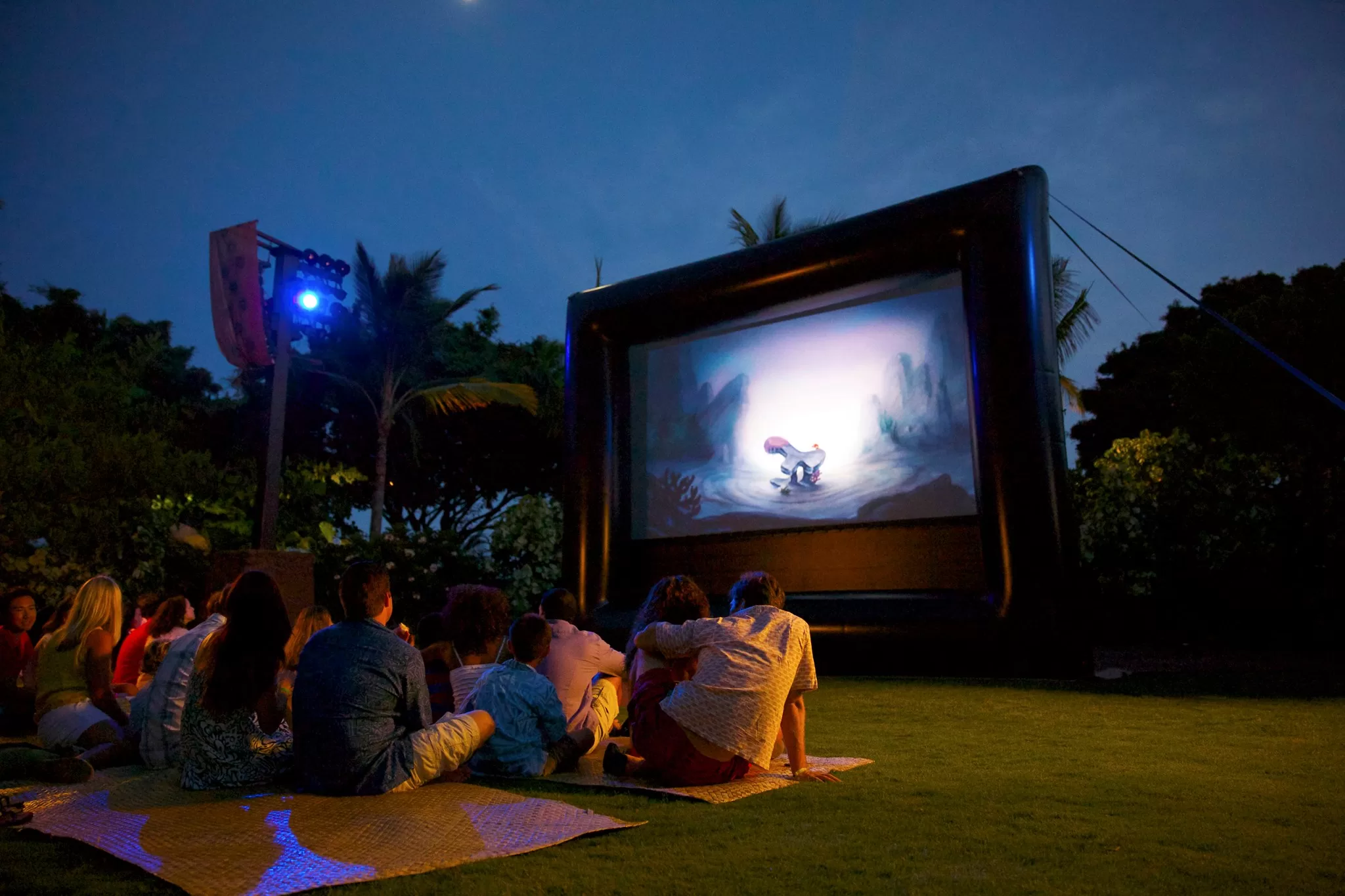 Outdoor movie screen with people sitting on mats on the ground watching